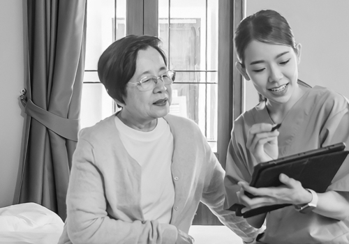 Woman Sitting On A Bed With A Nurse Who Is Reading Questions From A Tablet